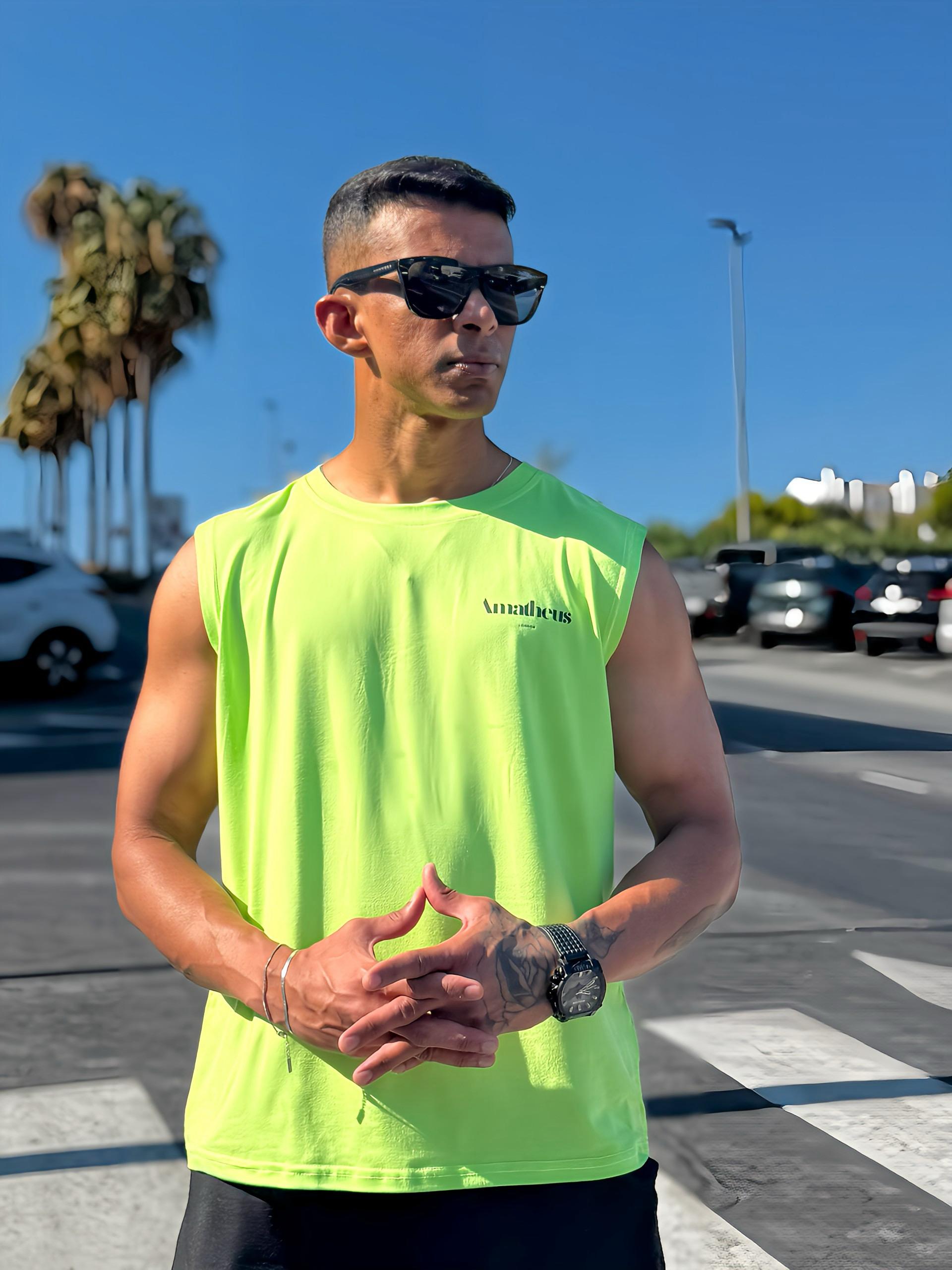 An athletic man wearing a neon green AMATHEUS Zenith Performance Tank and black sunglasses. He stands outdoors on a pedestrian crossing with hands clasped, a black watch visible on his wrist, and blurred cars, palm trees, and a clear blue sky in the background.