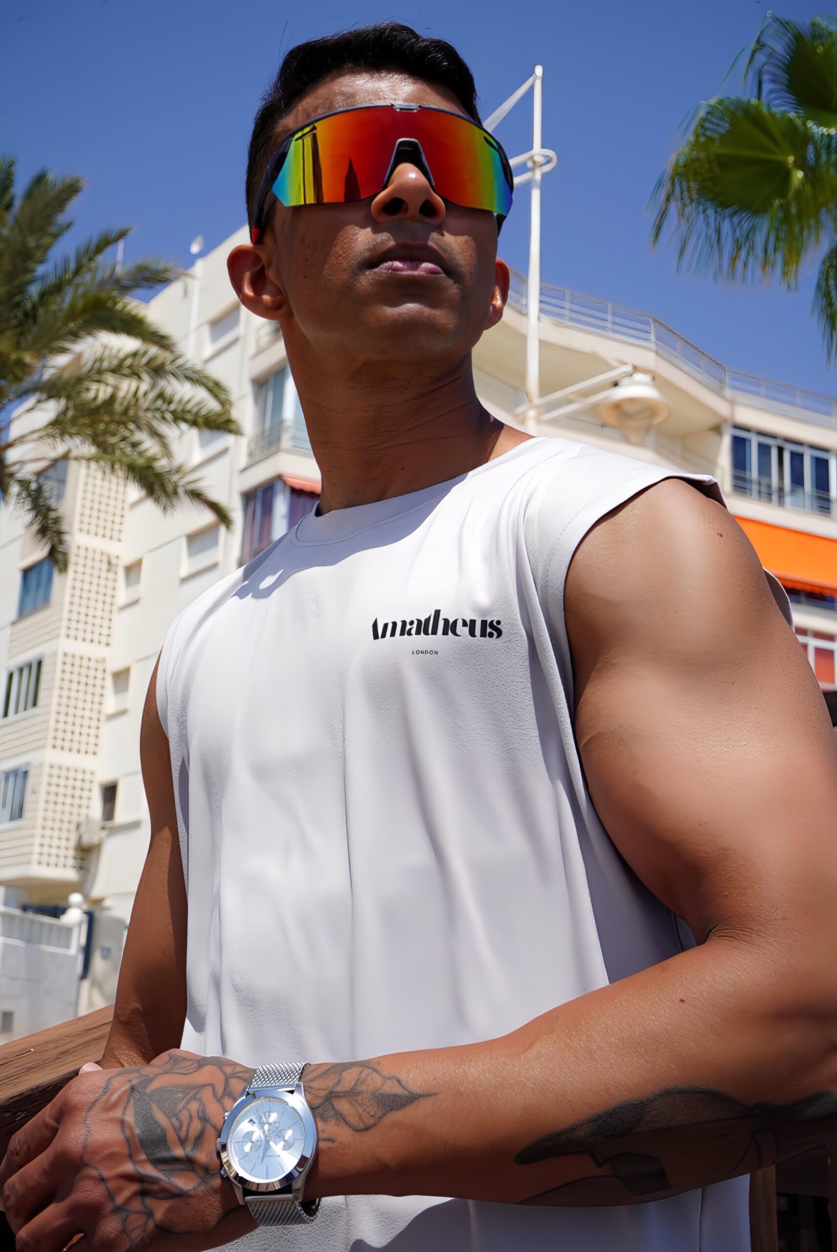 Hombre atlético con camiseta sin mangas gris claro AMATHEUS y gafas de sol deportivas de espejo, posando al aire libre en Benidorm con edificios blancos y palmeras de fondo bajo un cielo azul claro. El logo AMATHEUS en negro es visible en el pecho y lleva un reloj de pulsera plateado