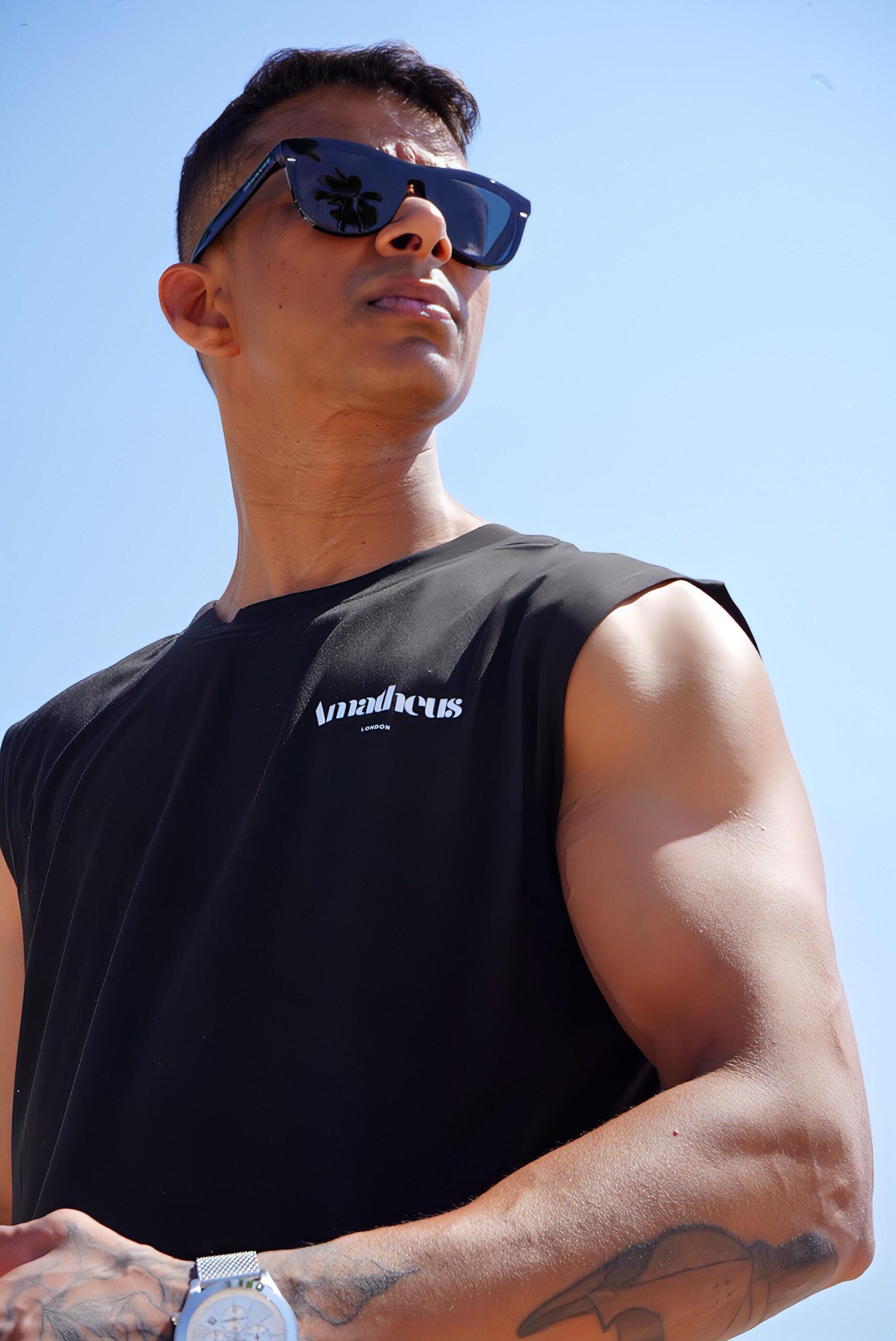 A person wearing a black AMATHEUS tank top, a silver chain, and a bracelet. The white 'AMATHEUS LONDON' logo is visible on the chest. The person is standing with hands clasped, in an indoor setting with a grey background.